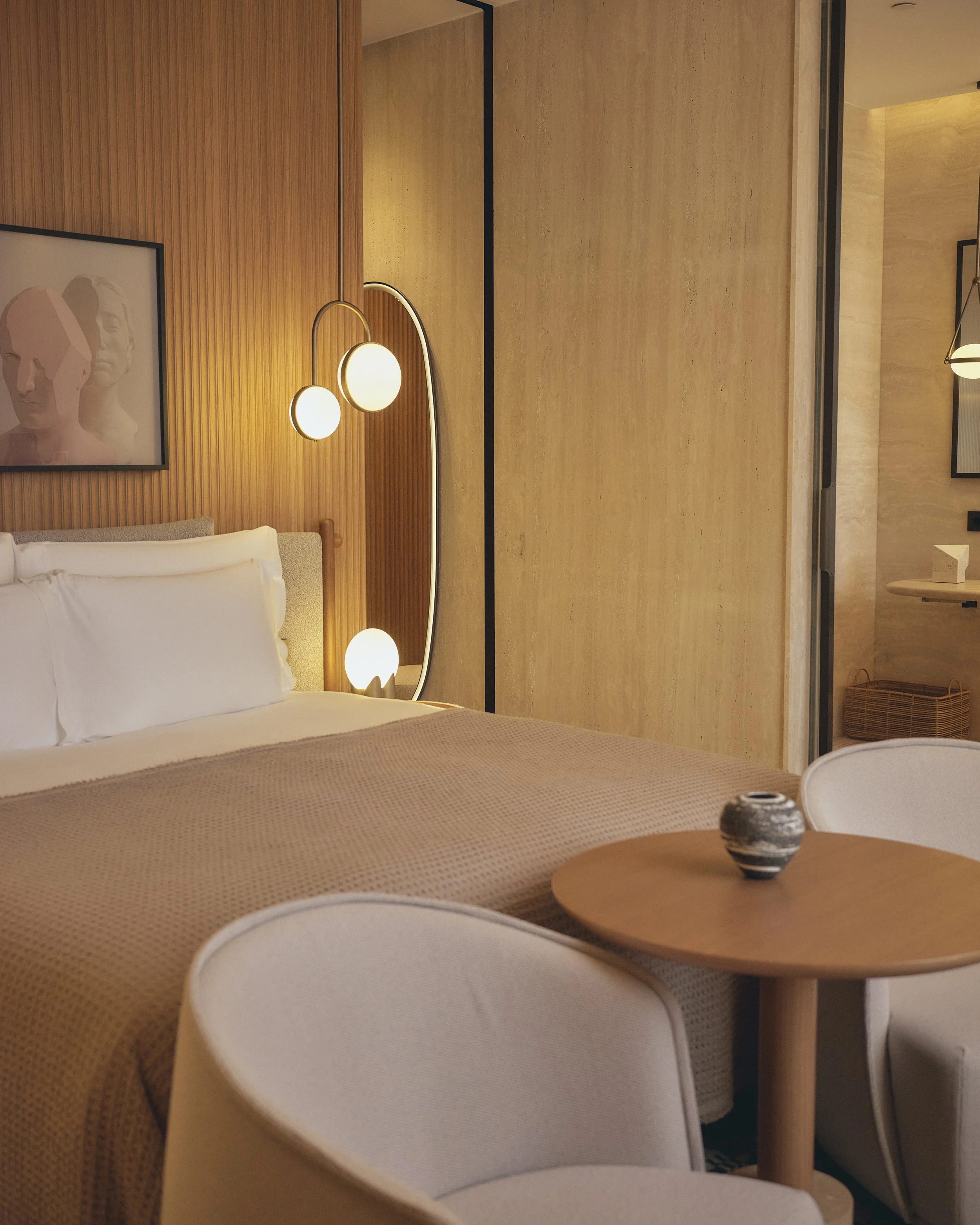 A cozy hotel room with a bed, a wooden wall with modern lighting, a round wooden table with a decorative bowl, and framed artwork on the wall.