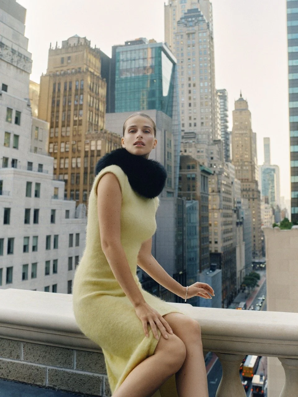 A woman sitting on a balcony ledge overlooking a cityscape with tall skyscrapers and streets below, with buildings in the background.