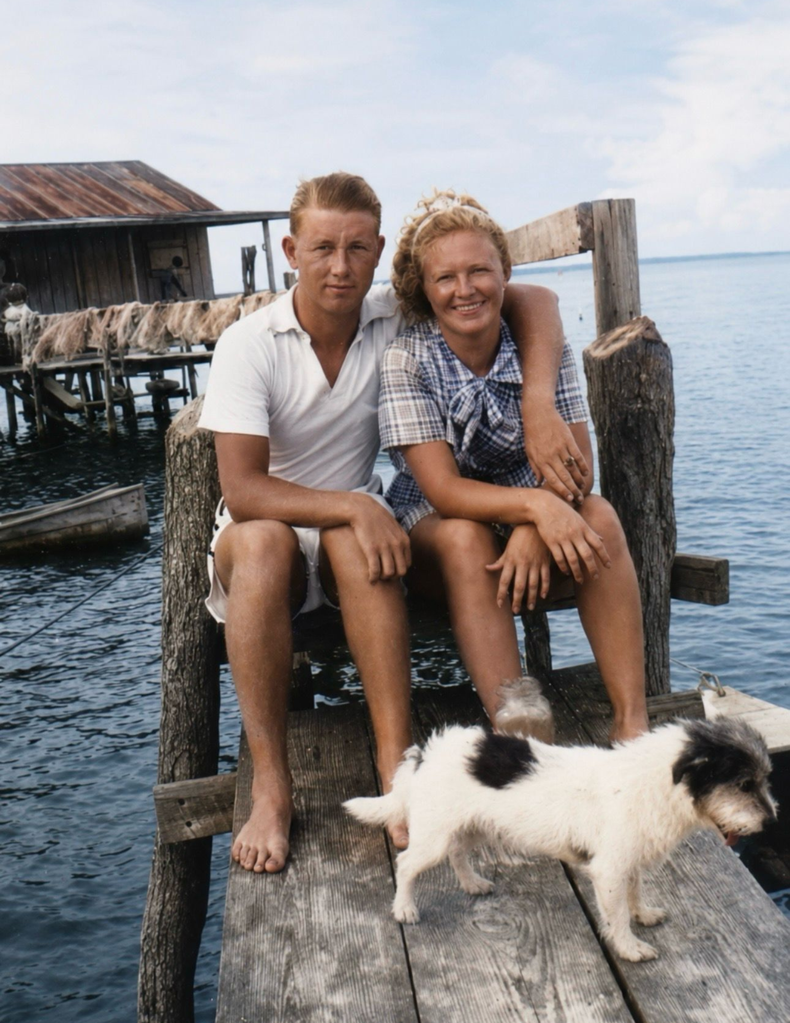 Smiling couple sits on a weathered seaside dock beside a small black-and-white dog, with rustic fishing huts and calm blue water behind them.