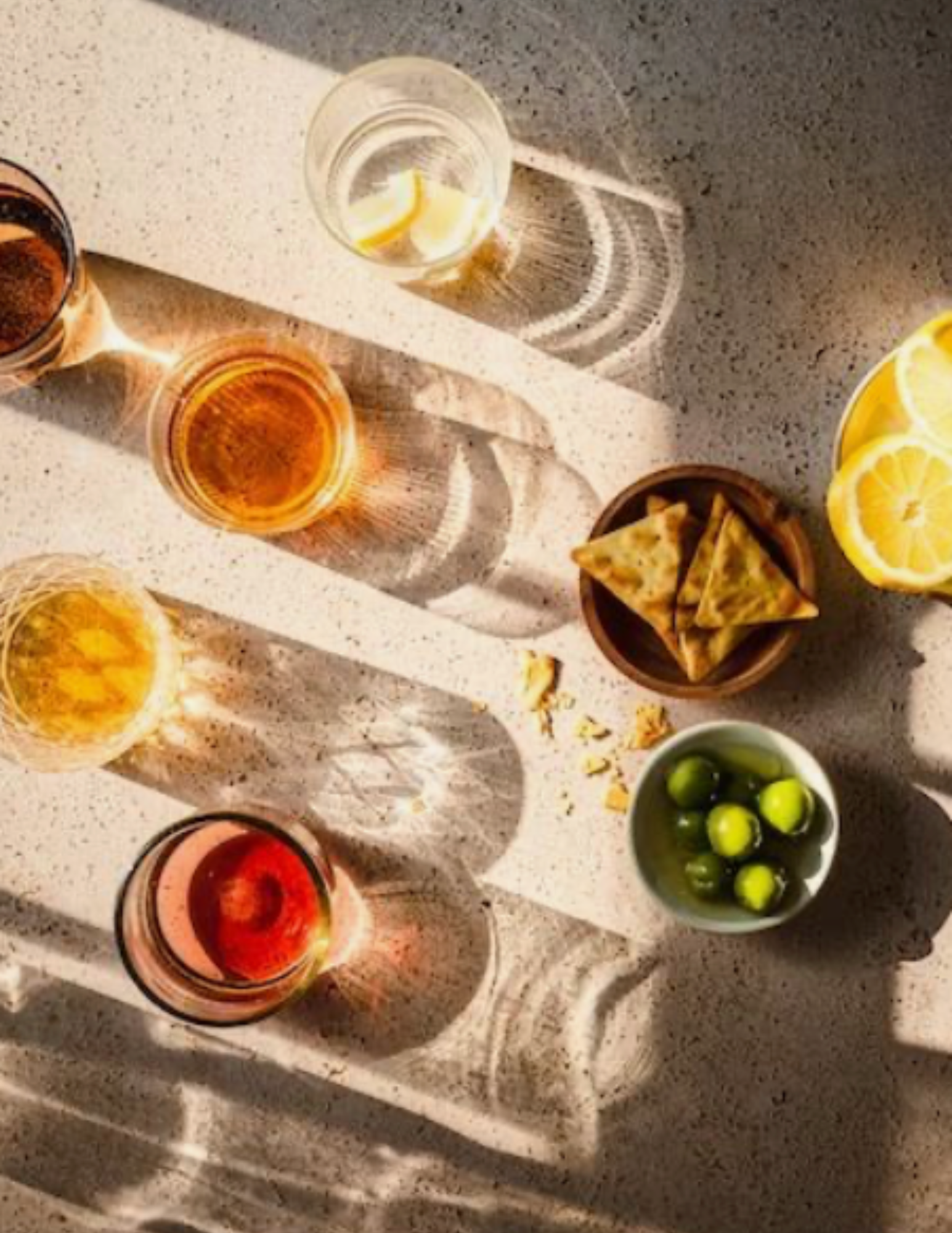 Sunlit table with glasses casting long shadows, containing drinks in warm hues. Nearby, lemon slices, green olives, and crackers create a relaxed ambiance.