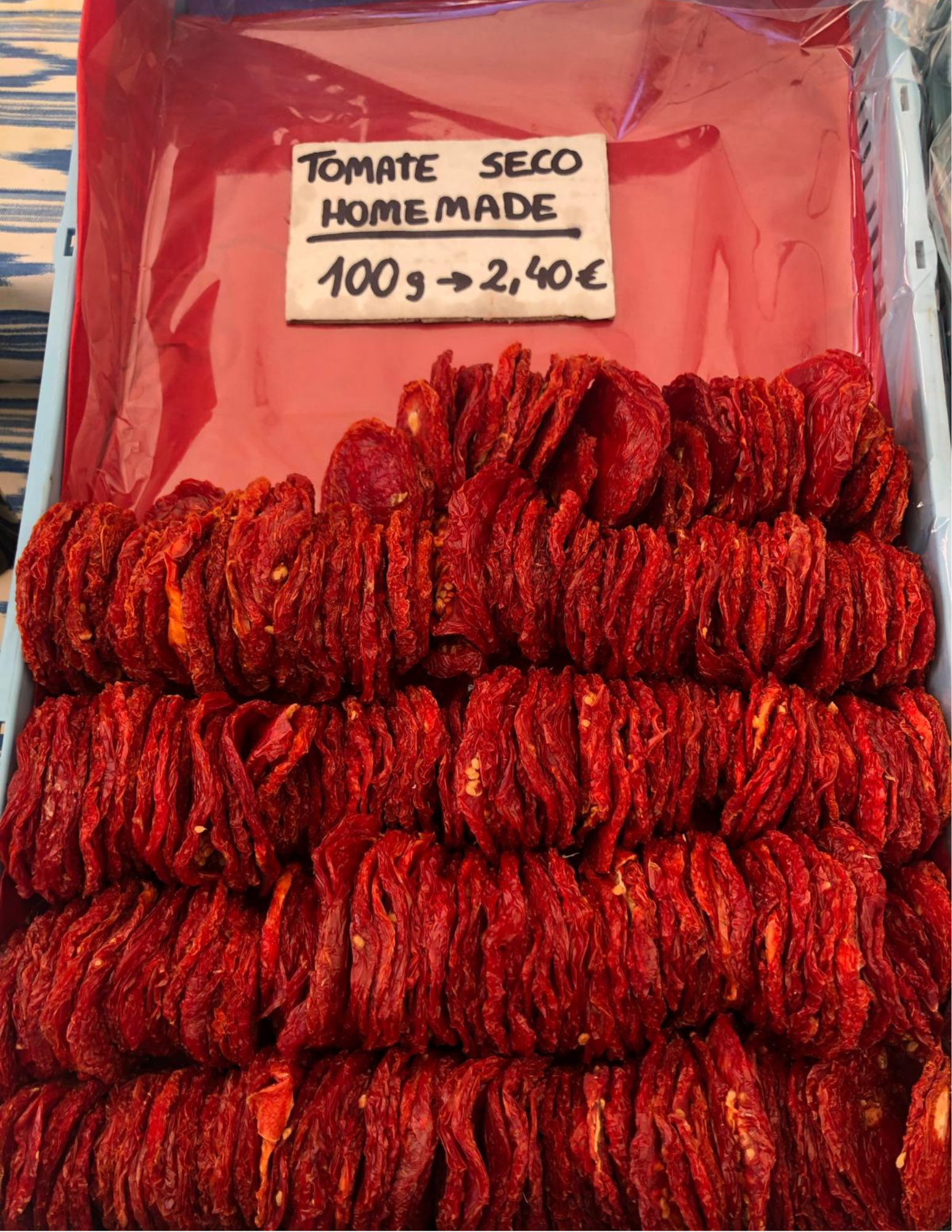 Stacks of dried red tomatoes are displayed neatly in rows. A sign above reads "Tomate Seco Homemade," priced at 2.40€ per 100g.