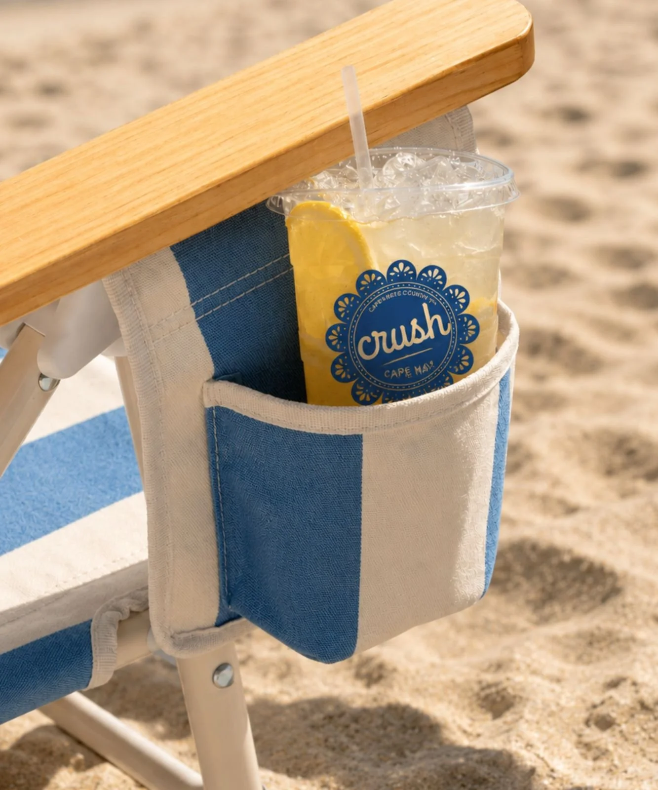 Iced lemon drink in a clear cup rests in a blue-and-cream beach chair cup holder on sandy shore, evoking a bright, relaxing day.