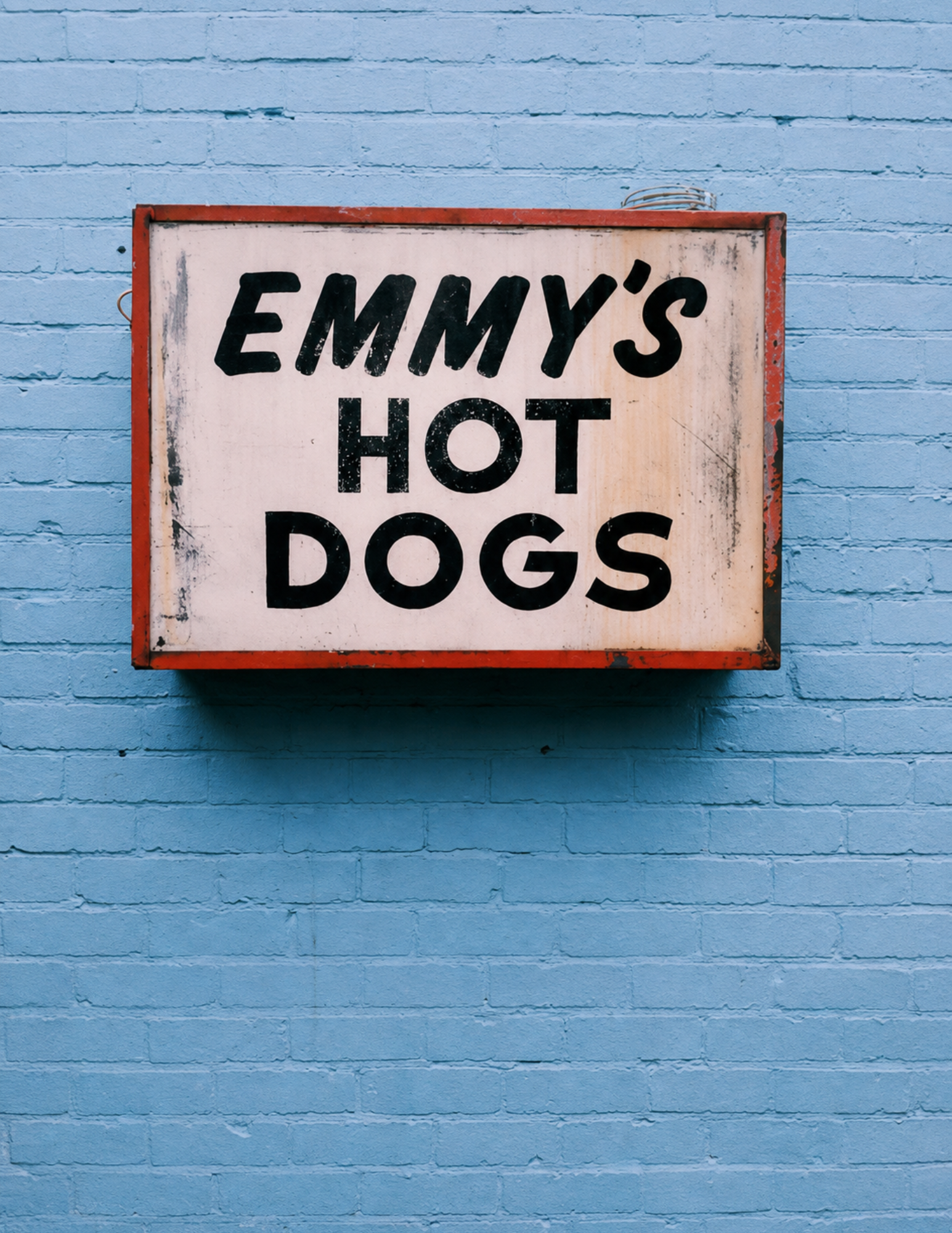 A retro sign reading "Emmy's Hot Dogs" is displayed against a blue brick wall. The red-bordered sign evokes a nostalgic, inviting feel.