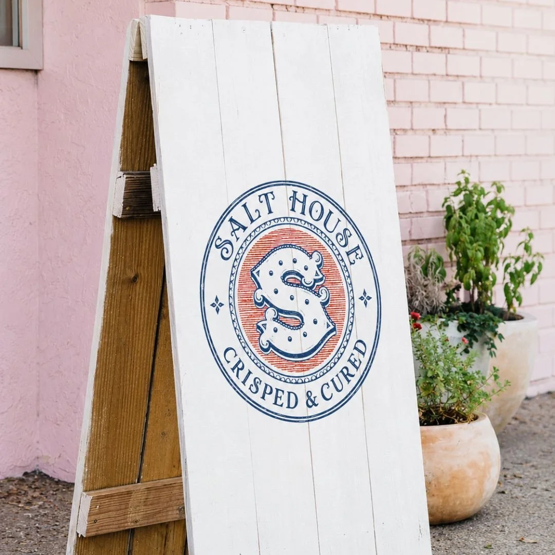 White sandwich-board sign reading “Salt House Crisps & Cutlery,” with a smiling salt-and-pepper logo, against a pink brick storefront.