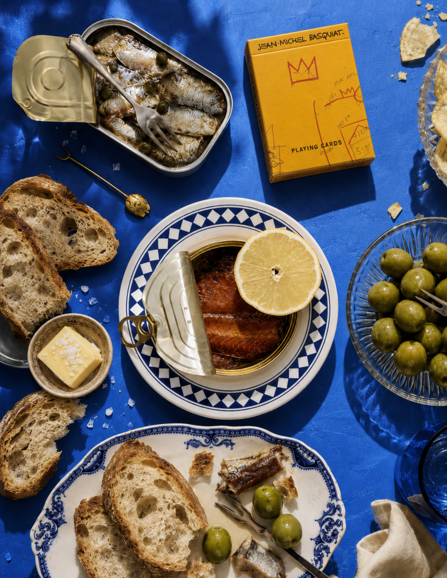 Tinned fish, olives, and crusty bread are artfully arranged on a vibrant blue table. A lemon slice and a yellow box of playing cards add a pop of color.
