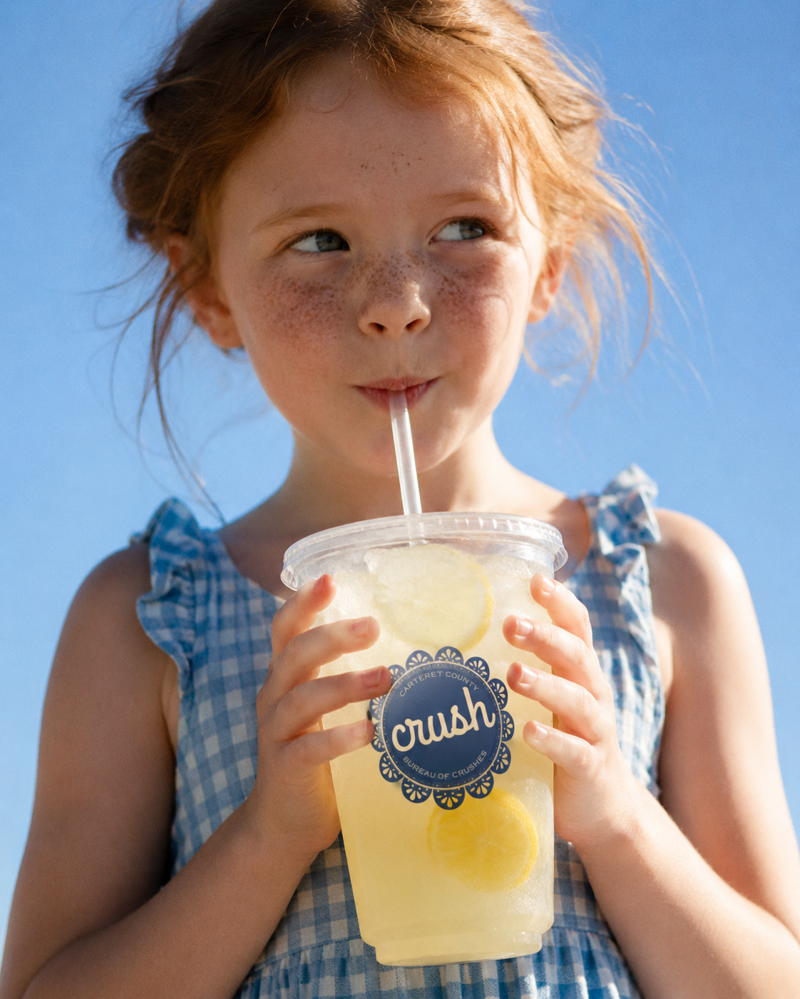 Freckled young girl in a blue gingham dress sips a lemon drink through a straw, holding the cup against a bright summer sky.