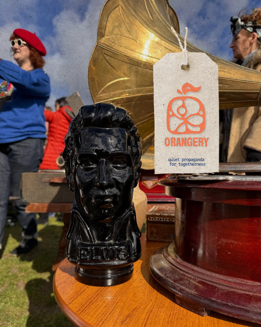 A black bust sculpture of a person's head on a wooden surface, with a brass gramophone, a paper tag reading 'ORANGERY' and 'quiet propaganda for togetherness', and people in the background at an outdoor event.