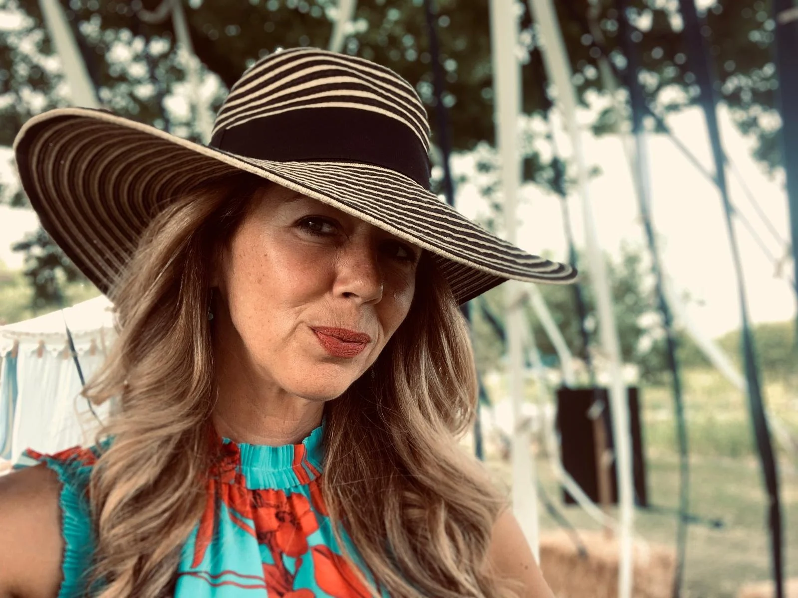 A woman with long wavy hair wearing a large striped floppy hat and a colorful sleeveless top, smiling subtly outdoors with trees and a metal structure in the background.