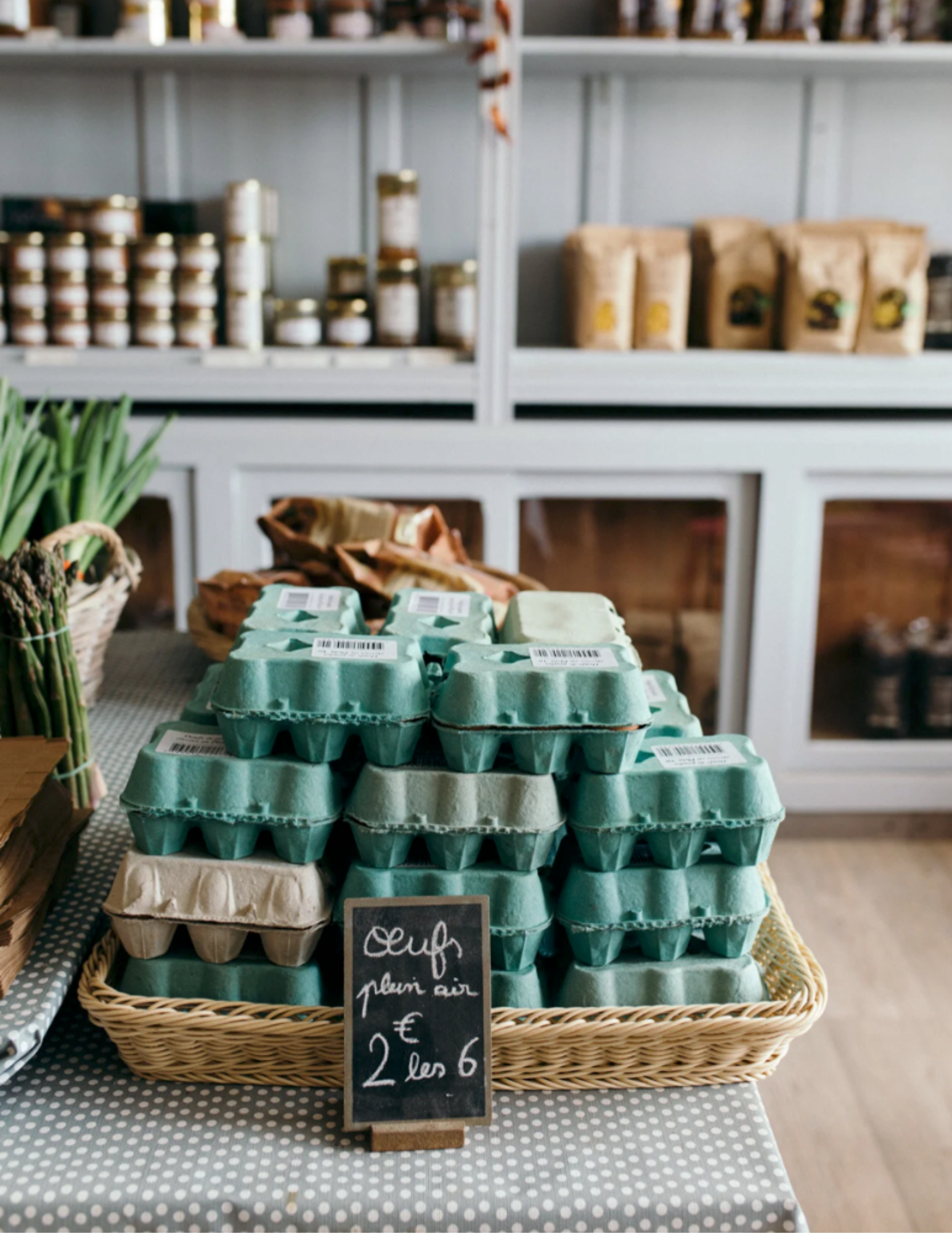 A display of green egg cartons stacked in a wicker basket on a checkered tablecloth, with a chalkboard sign reading "Oeuf plein air €2 les 6." Shelves with jars and bags are in the background.