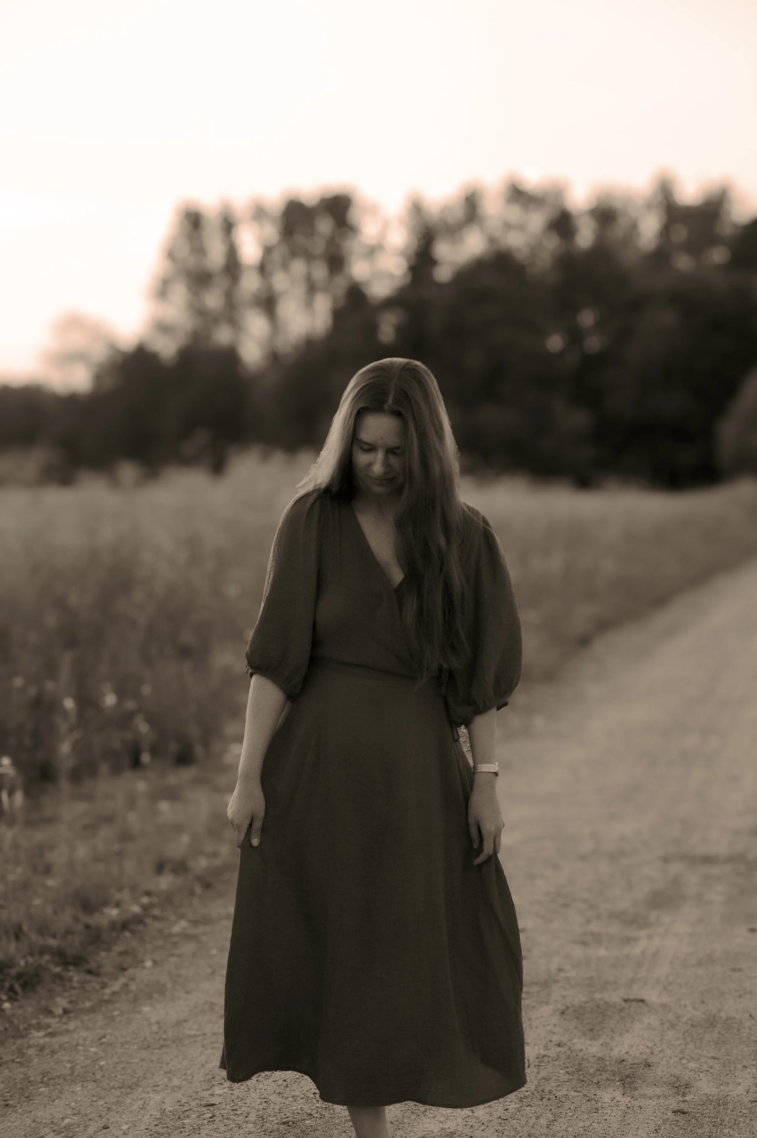 A woman with long hair wearing a dark dress standing on a dirt path outdoors during sunset, with trees in the background.
