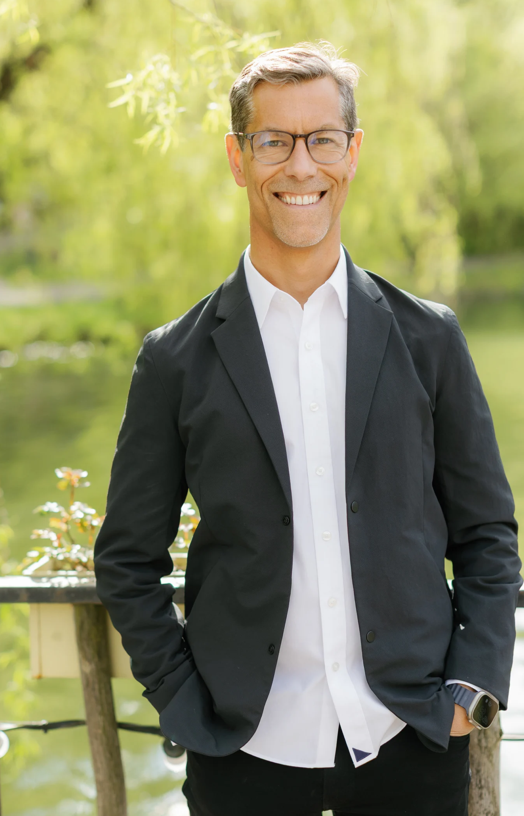 A smiling man in a black suit and white shirt, standing outdoors near a body of water, with trees and greenery in the background.