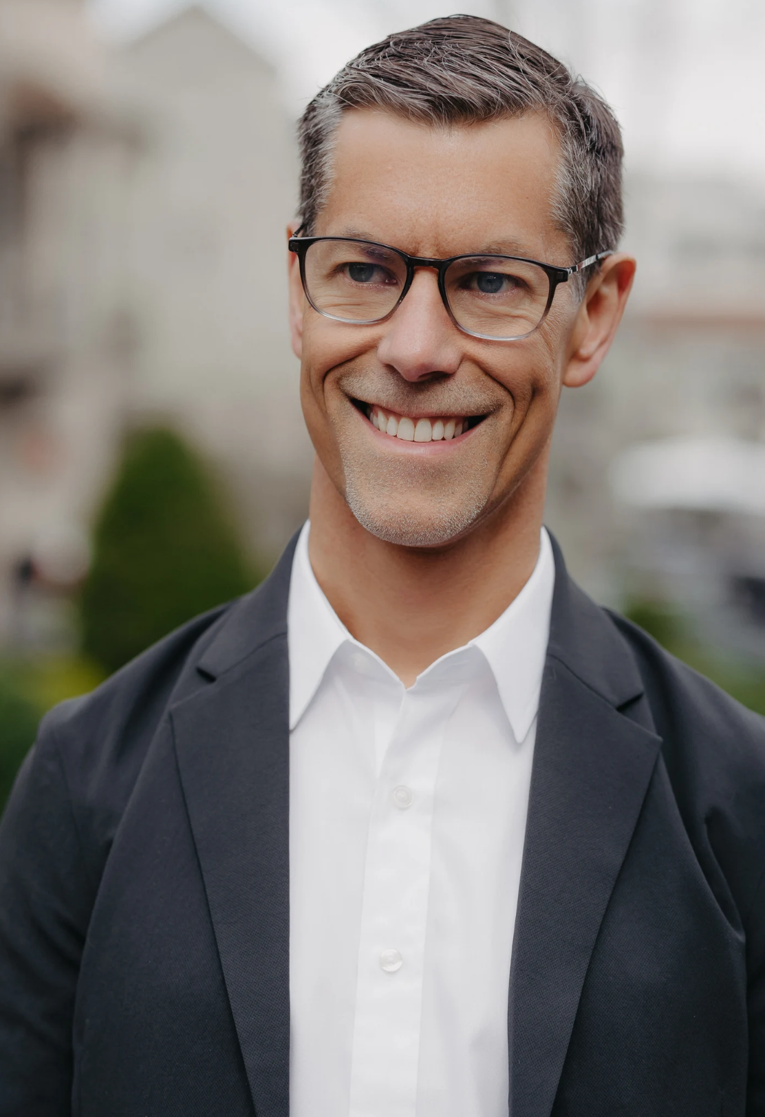 A middle-aged man with short gray hair, wearing glasses, a white shirt, and a dark blazer, smiling outdoors.
