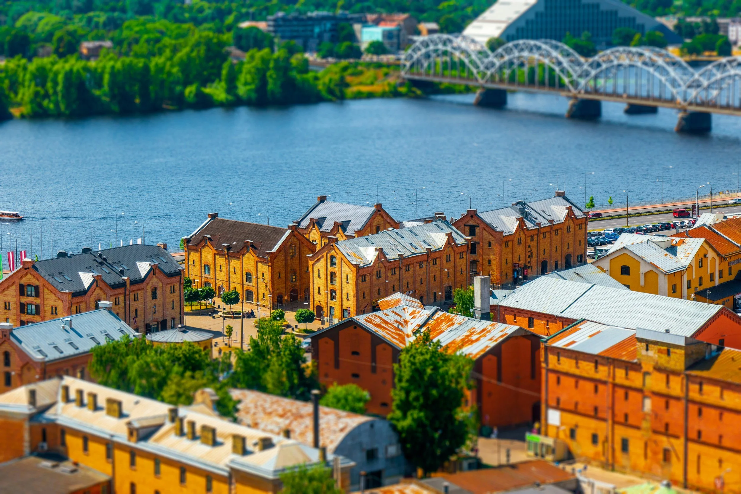 A cityscape of Riga, Latvia with colorful brick buildings along a river, a bridge with a domed roof structure, and lush green trees in the background.