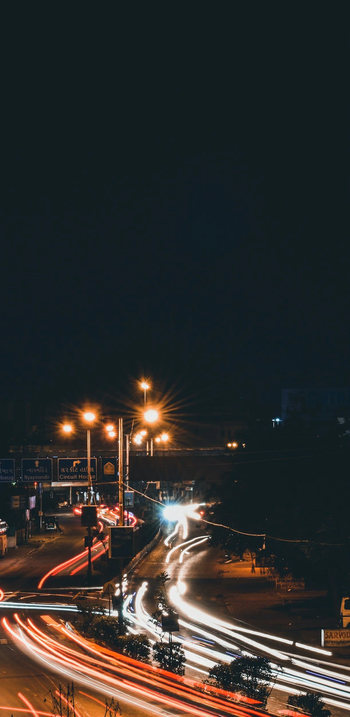 Nighttime city streets of Vadodara, India with light trails from moving vehicles, streetlights, and illuminated signs and billboards.