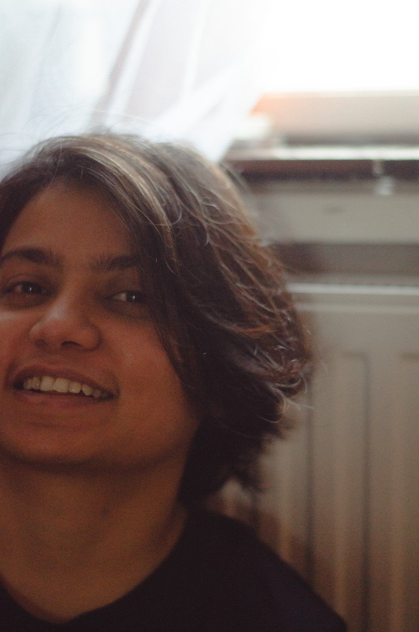 Photographer, smiling woman's face with brown curly hair and skin, wearing a dark top, background features blurred window blinds and a white wall.