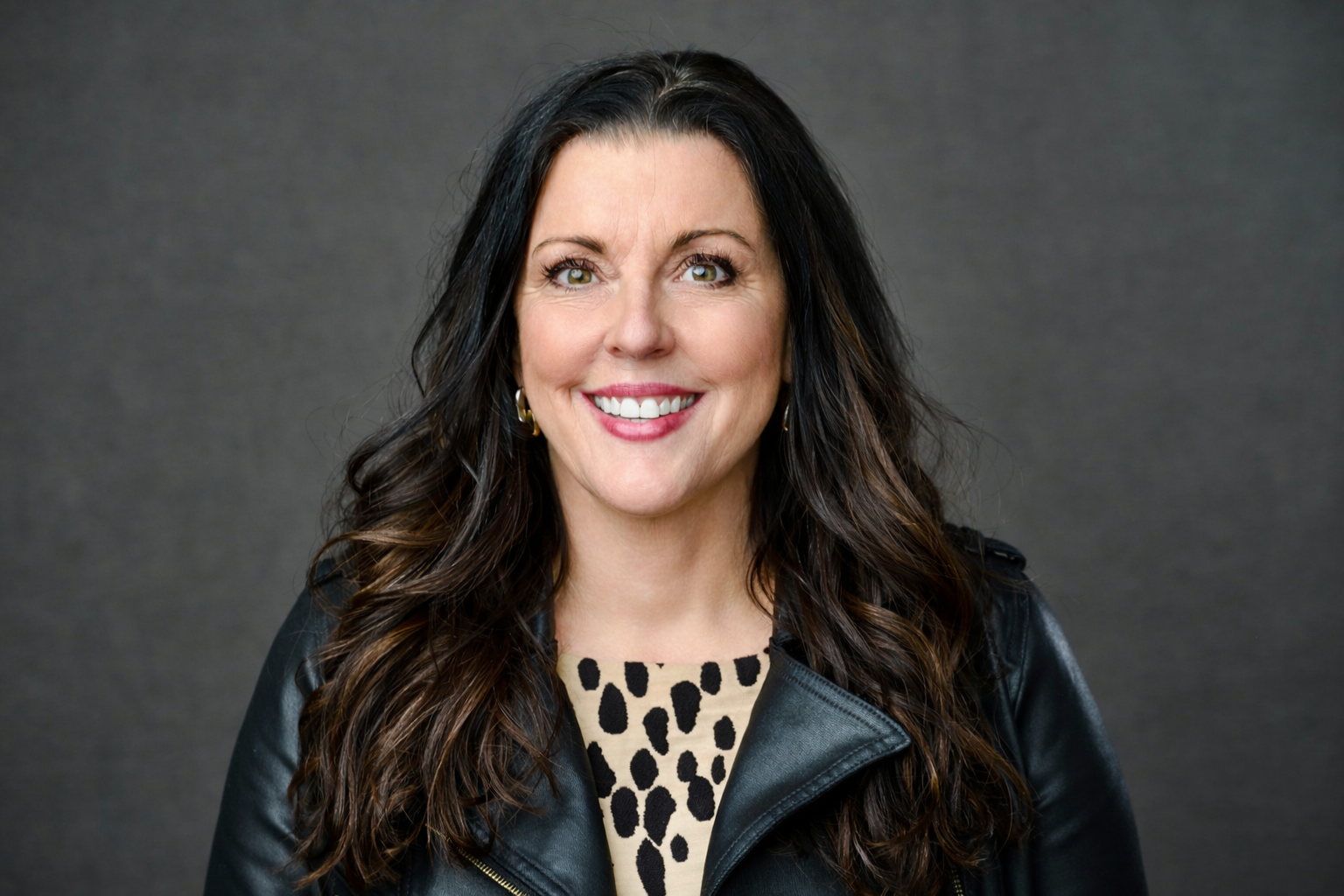 A woman with long dark wavy hair, green eyes, smiling, wearing a black leather jacket over a beige top with black spots, standing against a gray background.