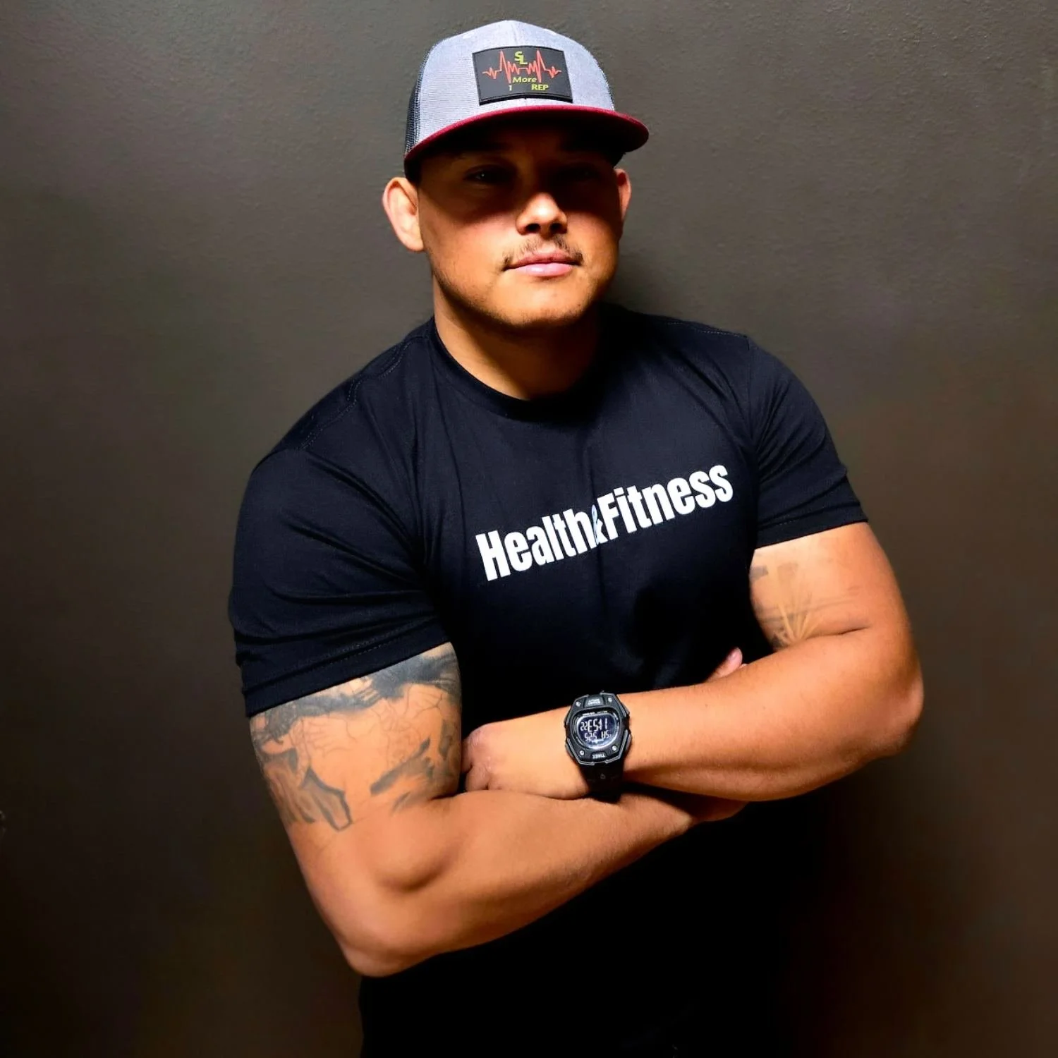A muscular young man with tattoos on his arm, wearing a black T-shirt that says 'Health&Fitness,' a gray and red cap, and a digital wristwatch, standing with arms crossed against a plain wall.