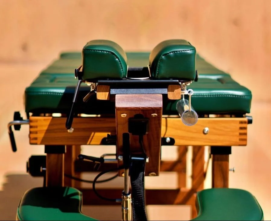 A close-up view of a chiropractic adjustment table with green padding and fitted wood frame, set against a wooden wall background.
