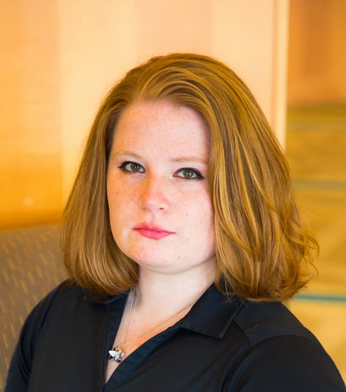 A woman with shoulder-length red hair, and light skin, wearing a black polo and a necklace, sitting in a room with warm yellow lighting.