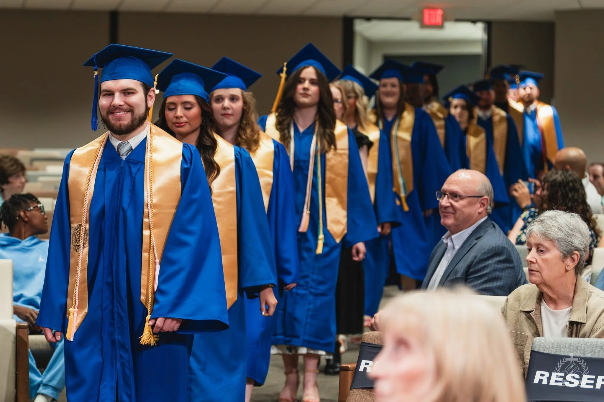 Group of diverse JSBC graduates in blue caps and gowns standing in line during a graduation ceremony.