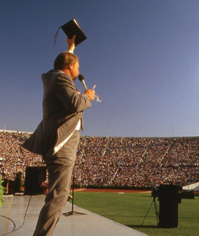 Jimmy Swaggart in a suit holding a microphone and raising a book in the air at a stadium with a large crowd.
