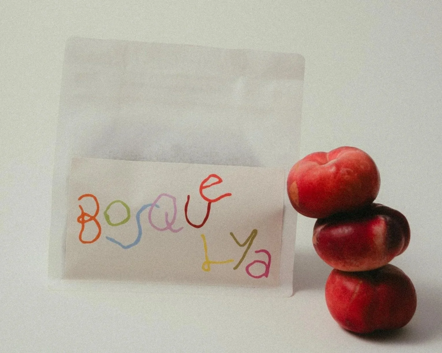 Product photograph of coffee bag, with a stack of four red peaches leaning on the right side of the bag. The coffee bag label reads 'BOSQUE LYA' in playful, colourful font. The background is plain white and lit with soft, warm light.