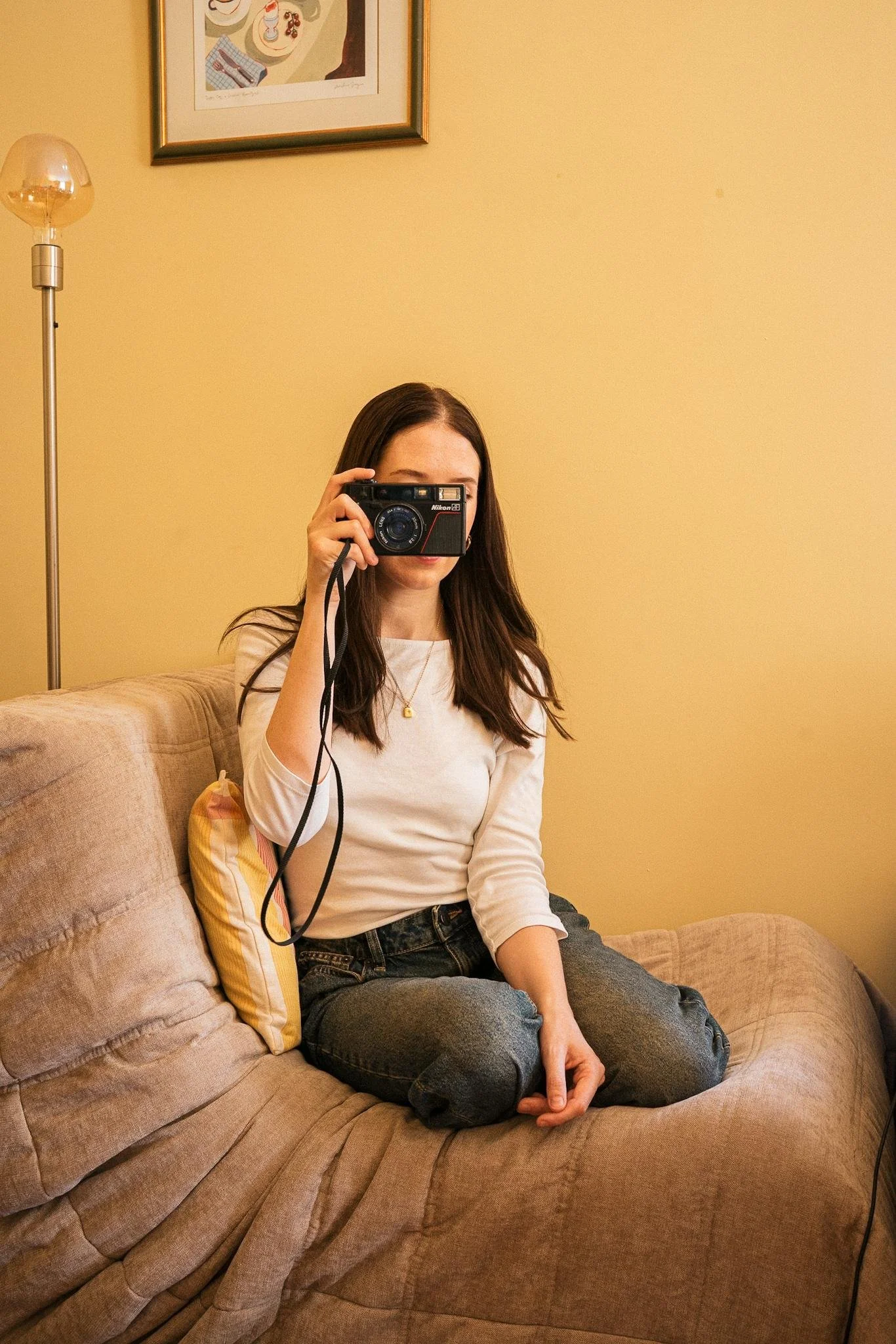 A woman with long brown hair sitting on a beige couch, holding a vintage film camera covering her face, wearing a white long-sleeve shirt and blue jeans. The background shows a yellow wall with a framed painting and a tall floor lamp.