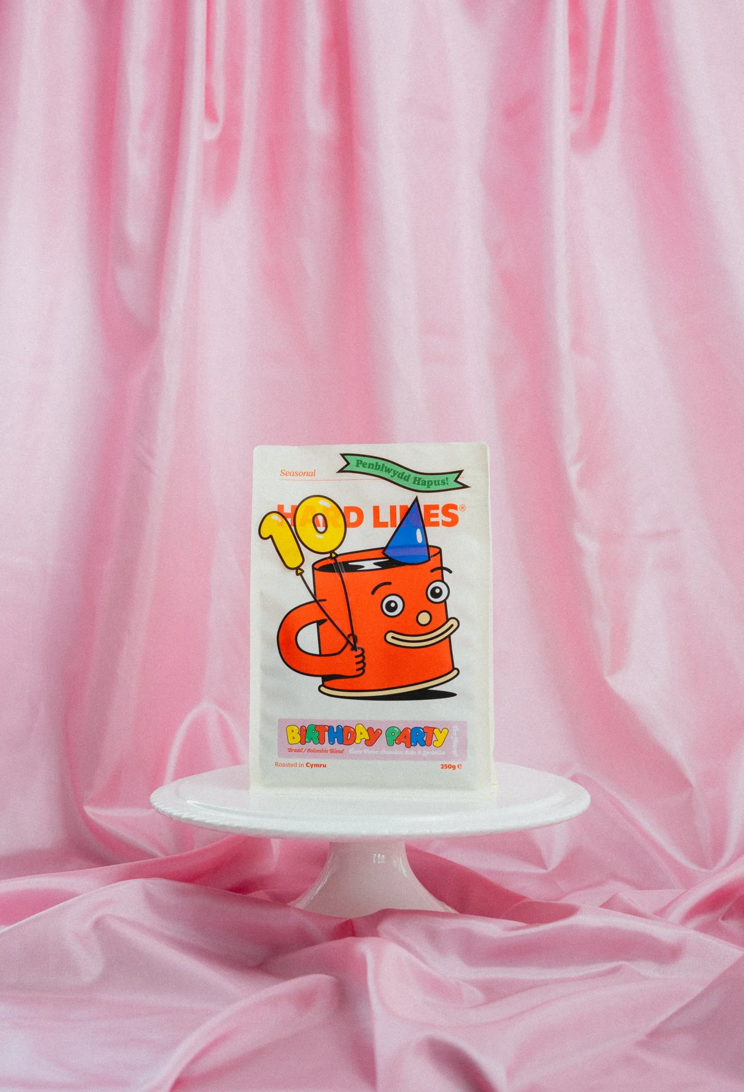 Pink birthday party backdrop with a white cake stand in front, a colourful coffee brand package featuring a smiling mug with a blue party hat sits on the stand.