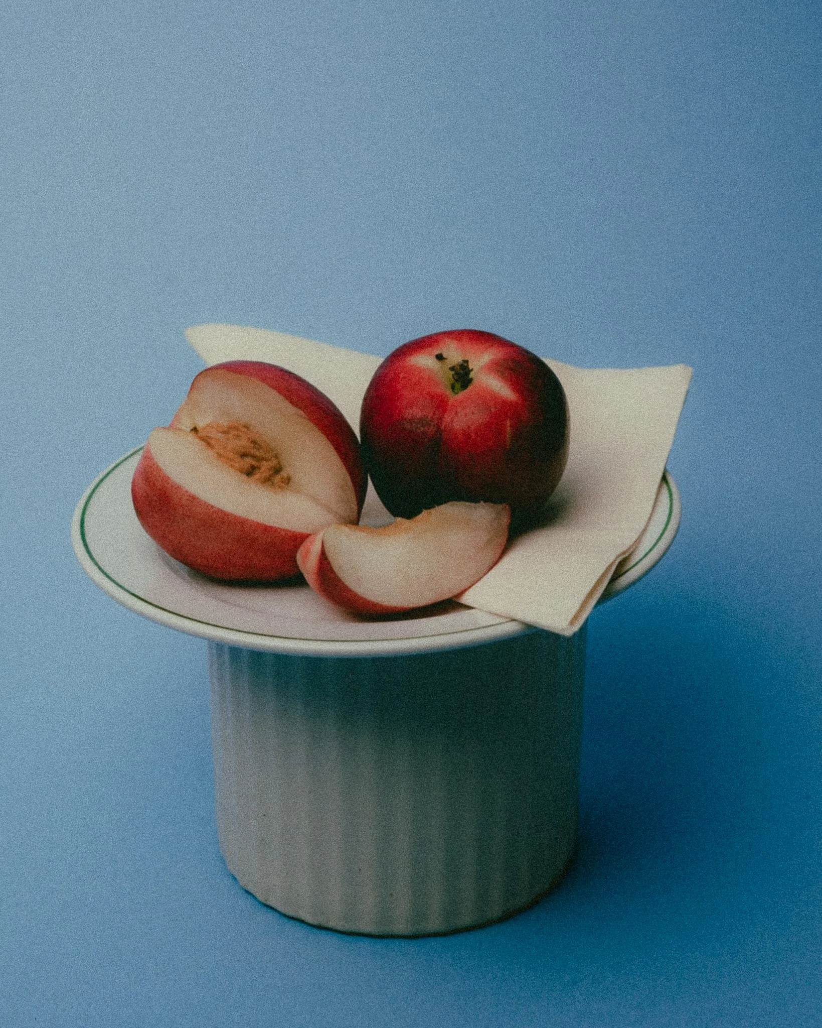 Still life product photograph of ceramic dish, containing two peaches, one whole and one sliced, on a folded napkin against a powder blue background. Lit with soft studio lighting.