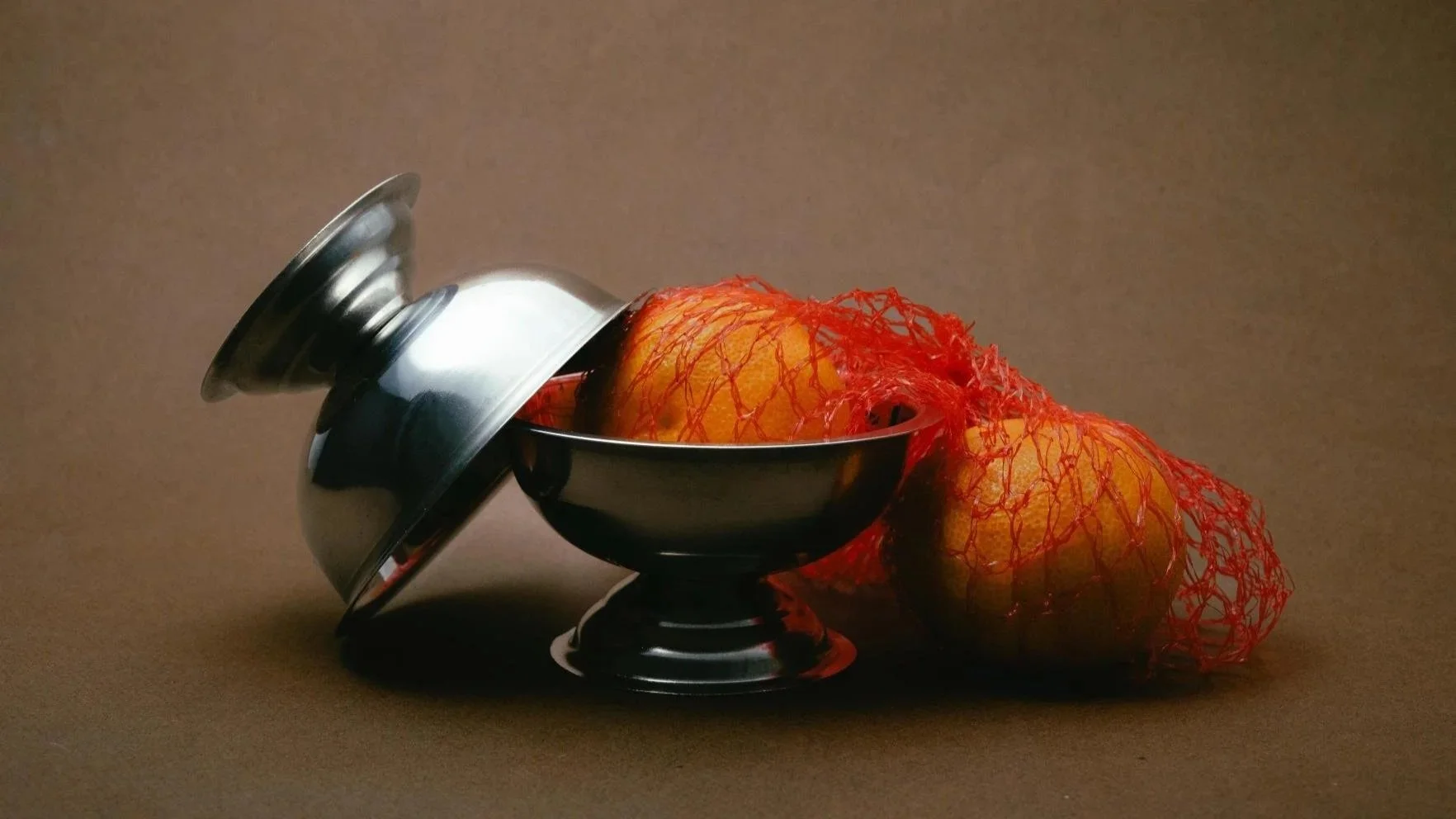 A black metallic punch bowl filled with peeled oranges covered with a red netted wrapper, with a matching lid tilted nearby, set against a plain brown background.