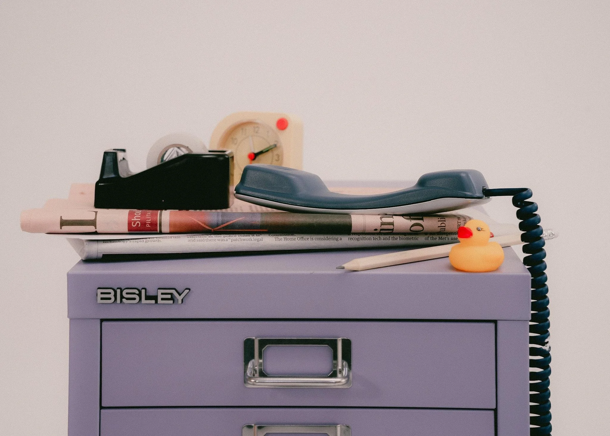 A purple medical storage cabinet with the label 'BISLEY' on the front. On top, there's a stack of newspapers, a yellow rubber duck with a red beak, a vintage black telephone with a rotary dial, a small beige clock, a white pen, and a black telephone receiver connected by a coiled cord. The background is plain light gray.