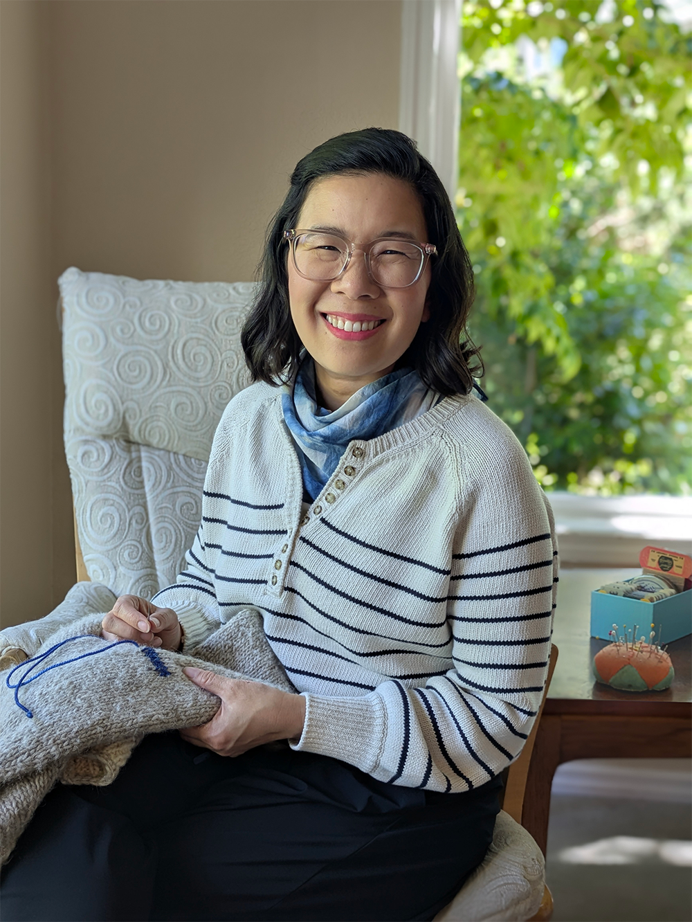 Photo of Charissa, smiling Asian American woman holding a needle and thread mending a sweater