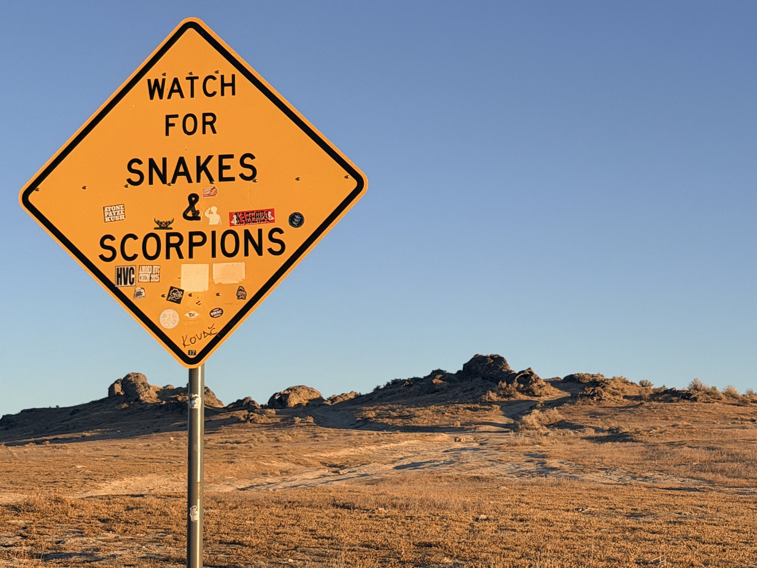 Orange diamond-shaped warning sign on a pole in a desert landscape, warning to watch for snakes and scorpions, with various stickers on it.