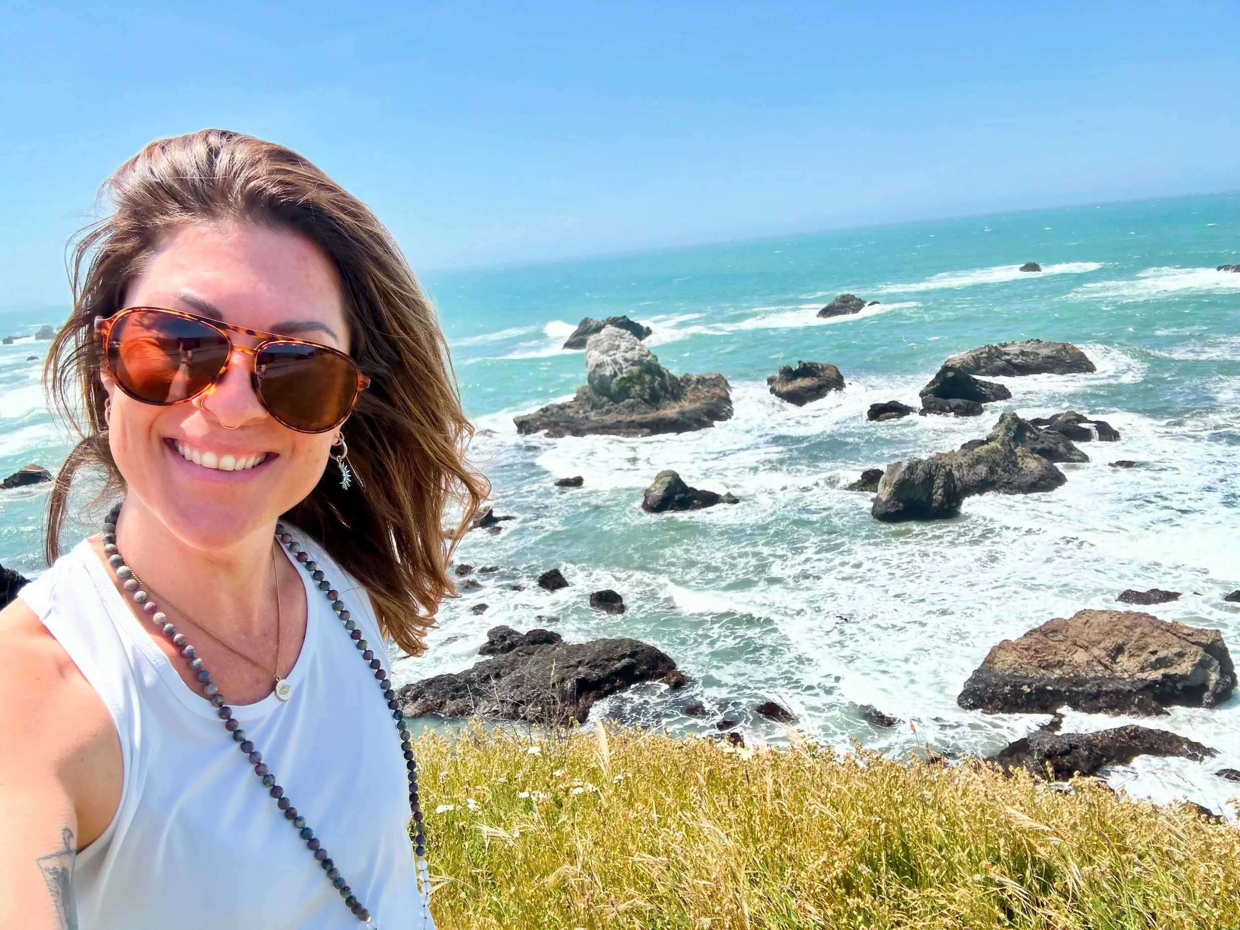 A woman with sunglasses and a beaded necklace smiling on a rocky shoreline with the ocean and a blue sky in the background.