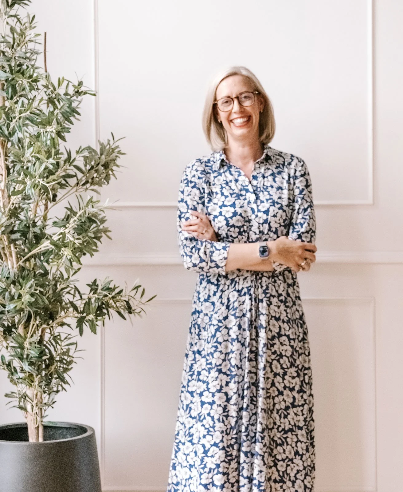 A woman with blonde hair, glasses, and a watch, smiling and wearing a long, floral dress, standing with her arms crossed in front of a white wall with paneling, next to a large potted plant.