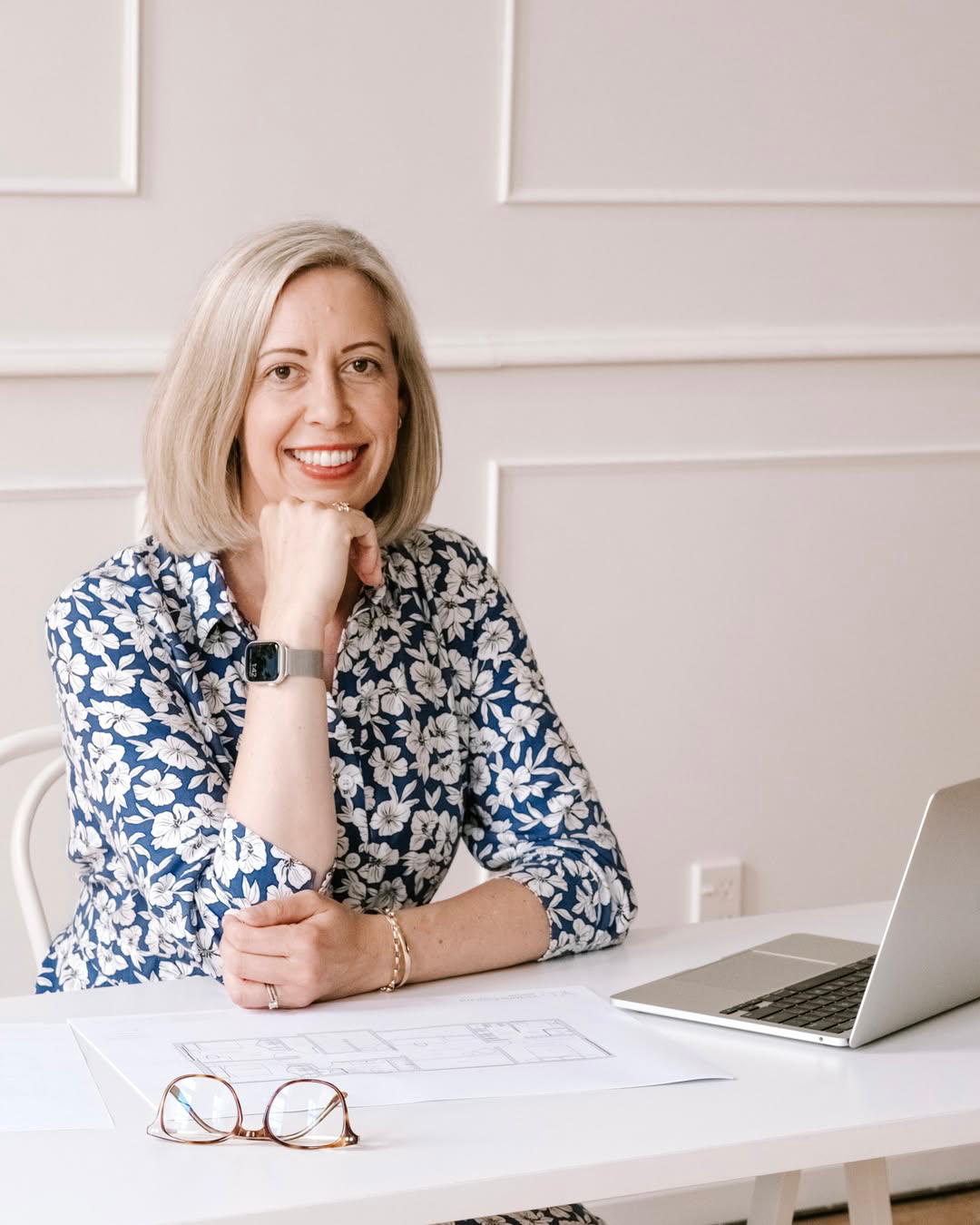 A woman with blonde hair wearing a blue and white floral shirt, sitting at a white desk with a laptop, eyeglasses, and architectural plans. She is smiling and resting her chin on her hand.