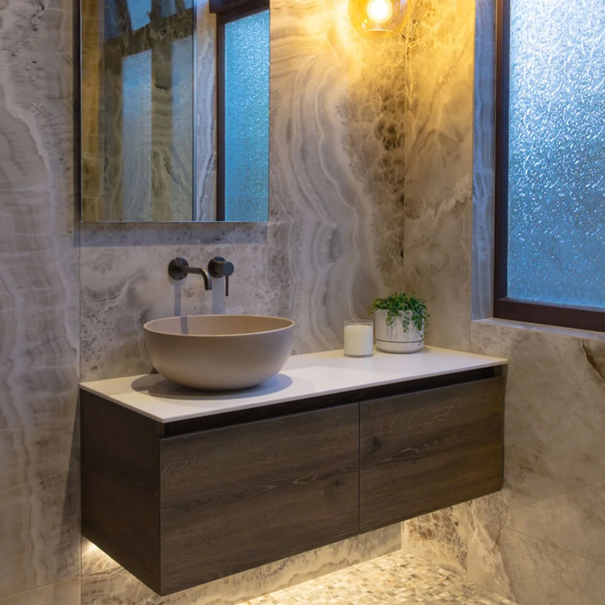 Modern bathroom with a white countertop, round beige vessel sink, wall-mounted faucet, marble walls, a mirror, frosted glass window, a candle, and a small green plant.