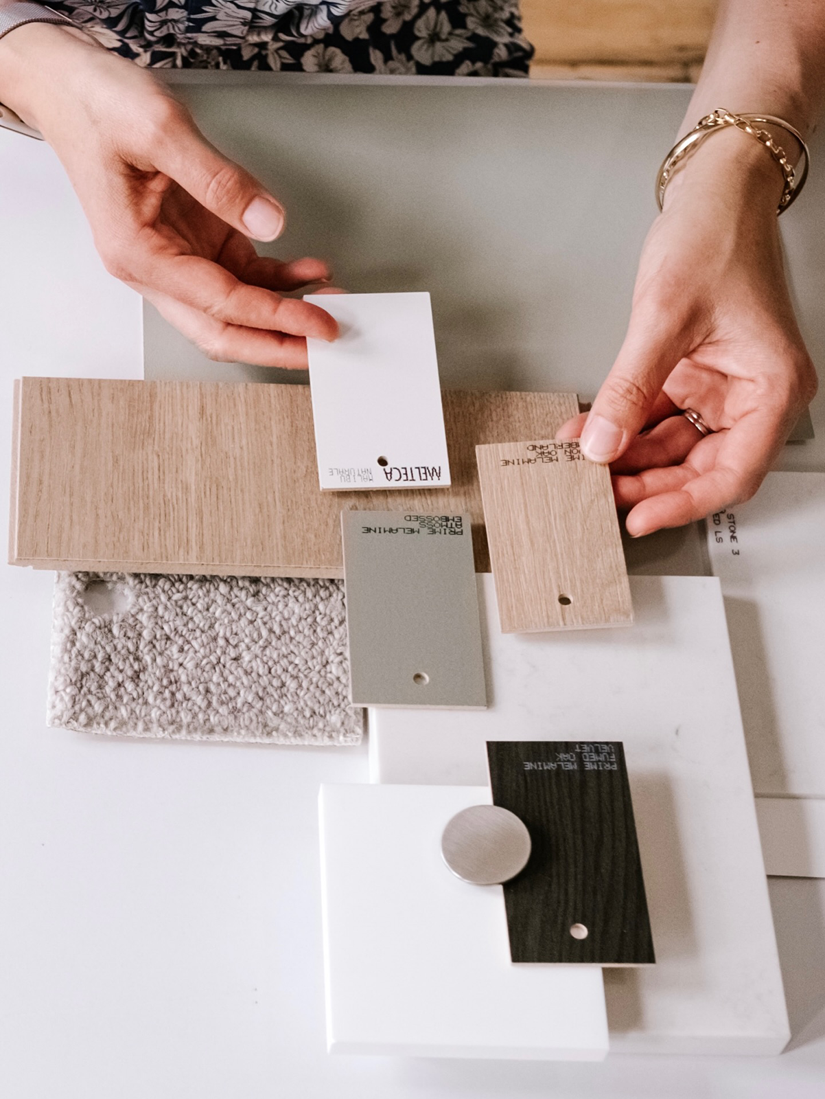 Hands holding sample materials including wood, paint, fabric, and tile swatches on a table.