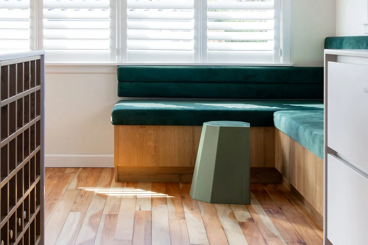 Interior of a room with wooden flooring, green cushioned built-in bench seating, white horizontal window blinds, and a small green side table.