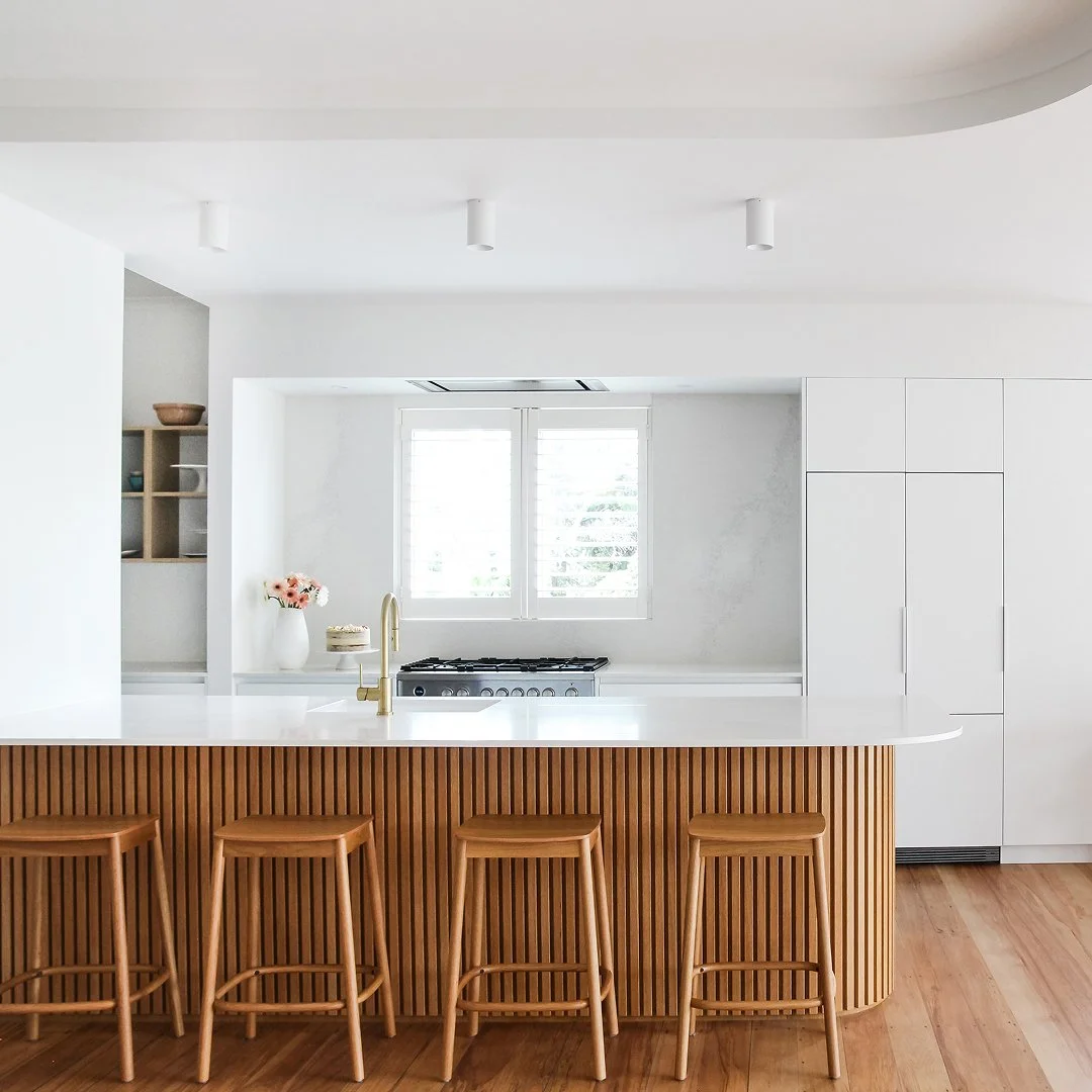 Modern kitchen with white cabinets, a white countertop island with wooden paneling, four wooden barstools, a gold faucet, a stove under a window with blinds, and wooden flooring.