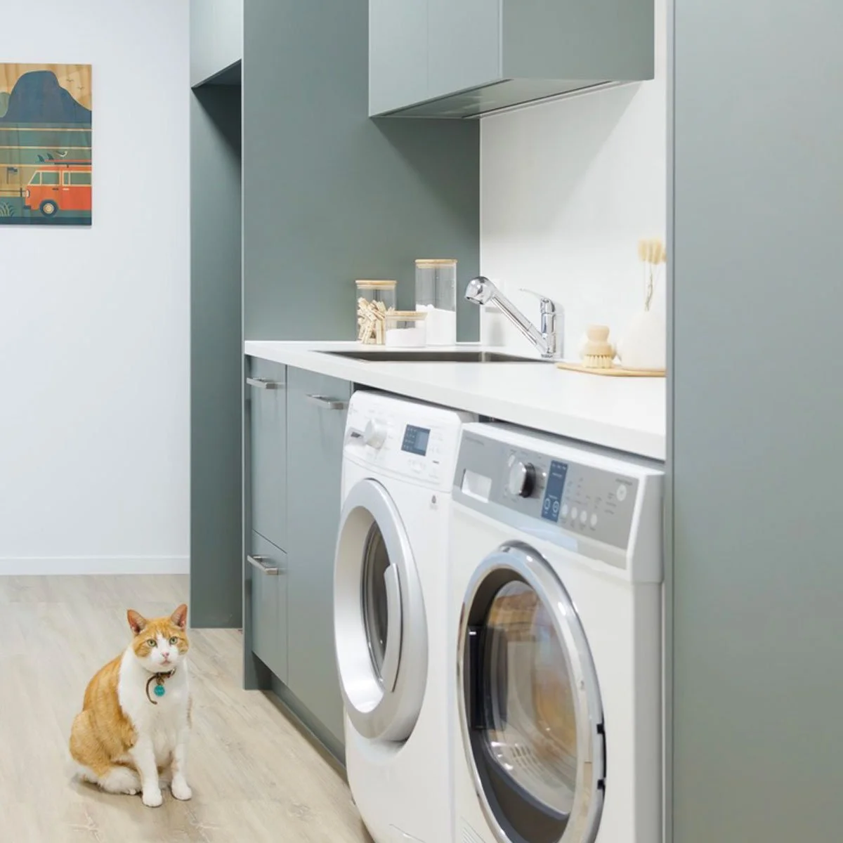 Laundry room with a cat sitting on light-colored wooden floor, grey cabinets, white countertop, washing machine, and dryer. Wall art and minimal decor.