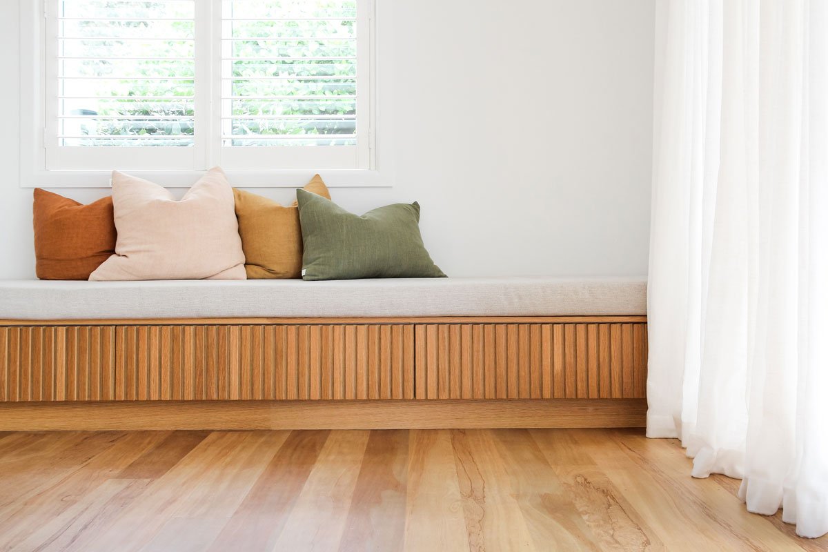 A minimalist sitting area with a wooden bench and five colorful cushions in front of a window with plantation shutters and white curtains on the side.