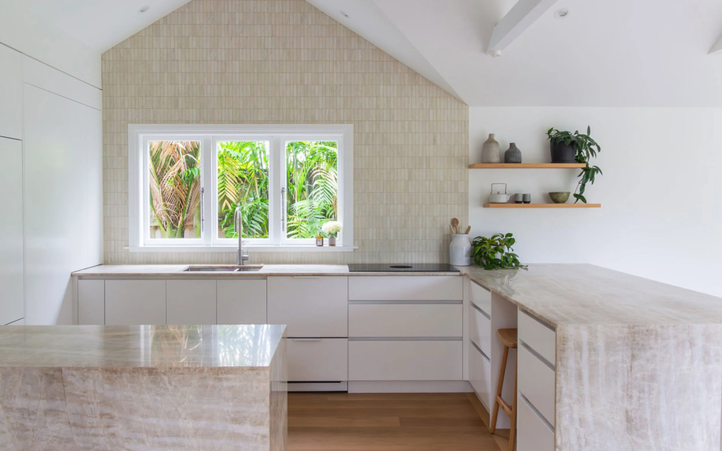 A modern kitchen with white cabinets, a beige marble countertop, a window above the sink showing lush green plants outside, and two floating wooden shelves with decorative items and plants.