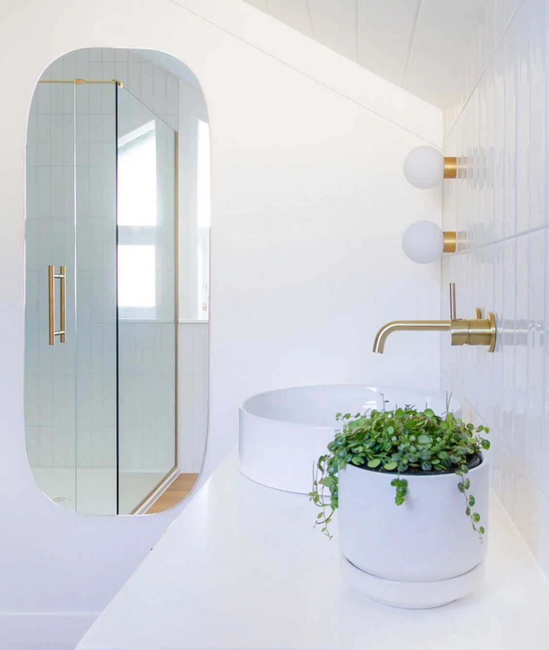 White modern bathroom with a round vessel sink, gold faucet, potted green plant, two spherical wall lights, and a mirror reflecting a window and glass shower door.