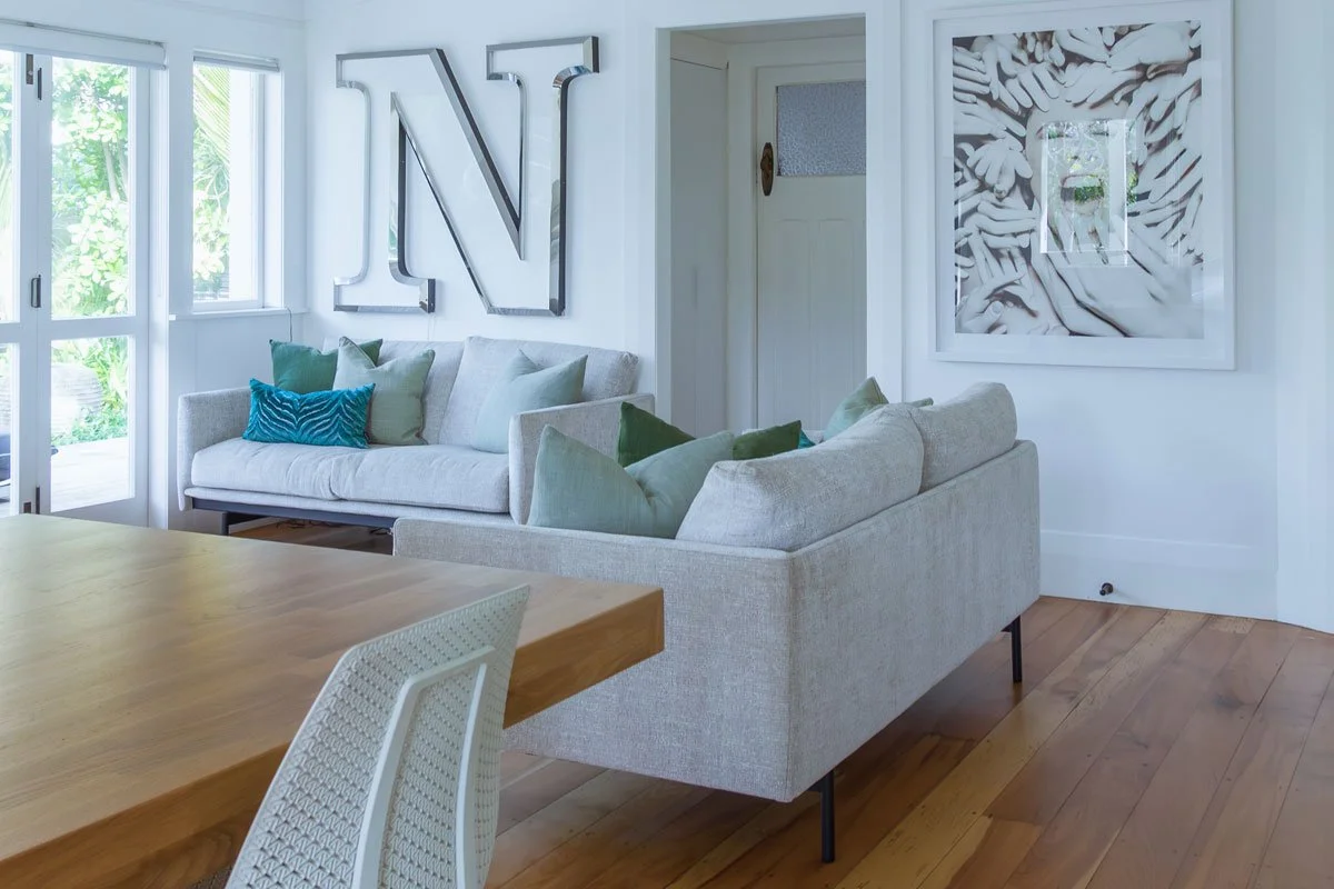 Living room with white walls, a light-colored sofa with multiple green and blue pillows, large window letting in natural light, wall art including a large metallic N, and decorative framed art with a swirling pattern.