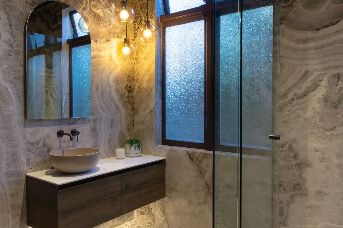 Modern bathroom with marble walls, a floating vanity with a vessel sink, a large mirror, a frosted glass window, and three hanging pendant lights.