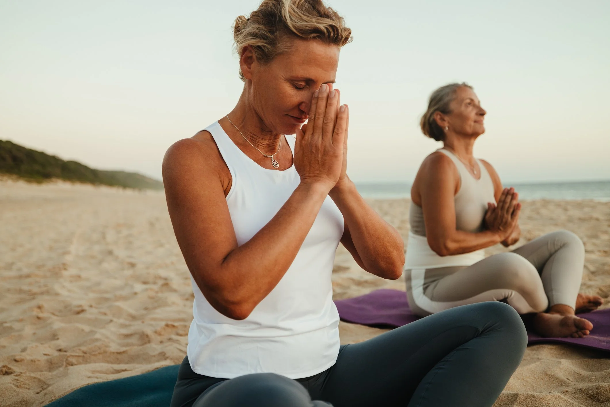 Nervous system regulation and midlife stress management coaching: Two women practicing yoga and meditation on a peaceful beach, with one woman in a white top holding her hands in prayer position against her forehead for mindfulness.