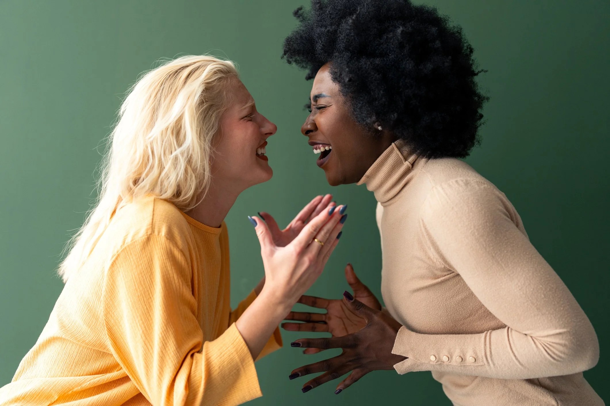 Menopause strategist support and consultation image: A blonde woman in a yellow top and a woman with curly black hair in a beige top laughing together closely against a green background, representing community, midlife coaching support, empowerment