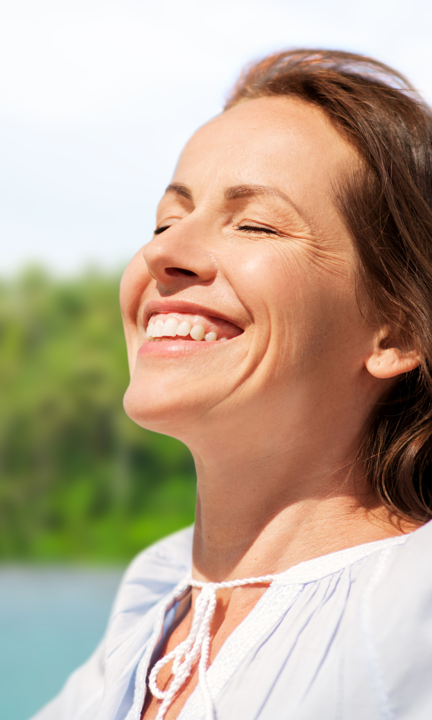 A vibrant woman smiling with her eyes closed as she enjoys the sunshine, representing total cortisol reduction, balanced hormones, and stress-free midlife living.