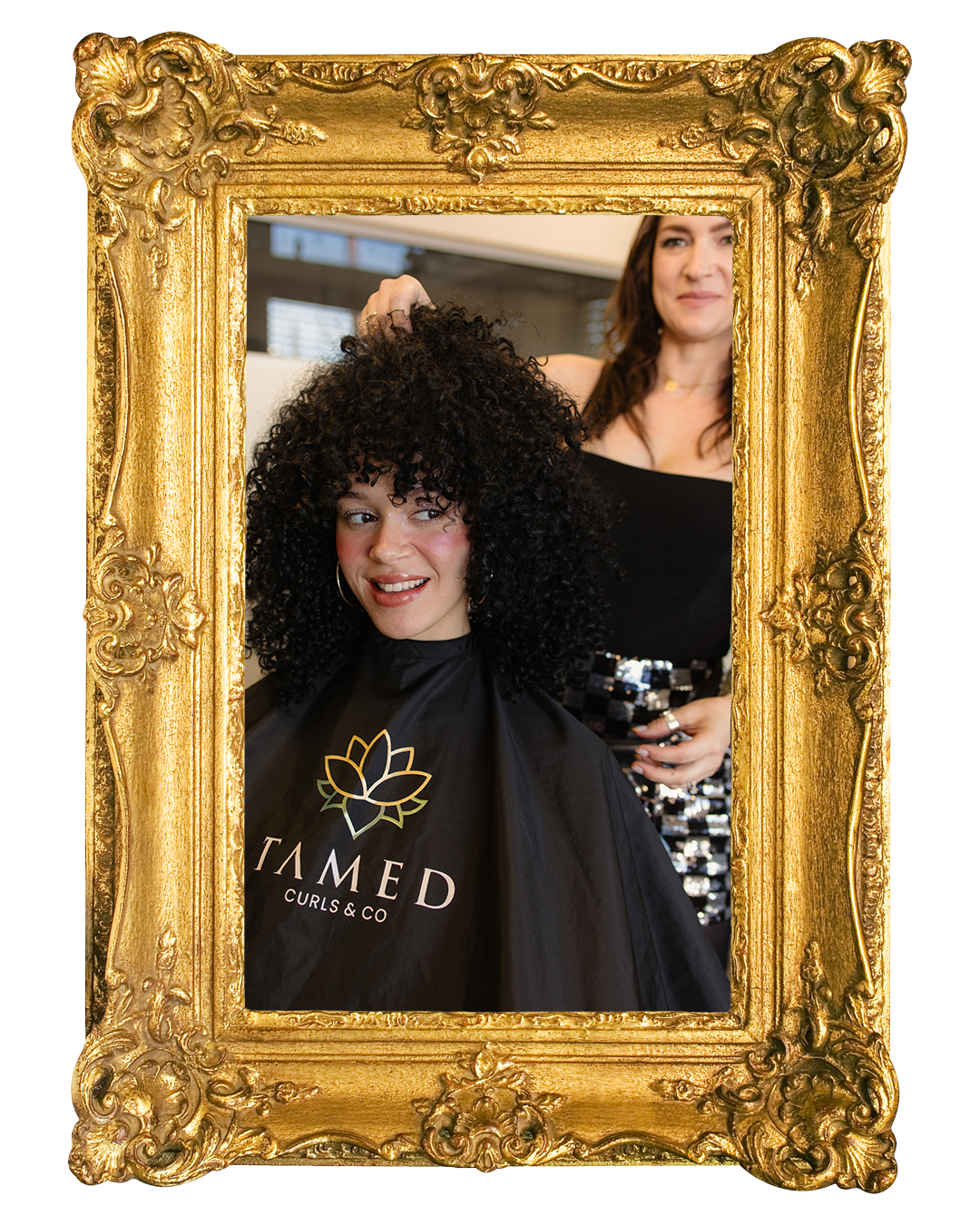 A woman with curly black hair sitting in a salon chair, smiling, while a hairstylist styles her hair. The woman is wearing a black cape with a lotus flower logo and the words "TAMED CURLS & CO." A large, ornate gold picture frame surrounds the scene.